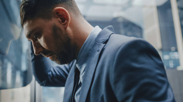 gestionnaire stressé descendant du bureau dans un ascenseur en verre dans un centre d’affaires moderne. spécialiste dévasté se sentant en colère après une réunion d’affaires infructueuse, se cognant la tête contre un mur. - banging your head against a wall photos et images de collection