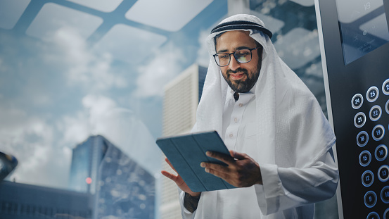 Exitoso hombre de negocios musulmán en kandura blanco tradicional montando ascensor de vidrio a la oficina en el centro de negocios moderno. Hombre usando tablet pc. Concepto de empresario saudí, emiratí, árabe. photo