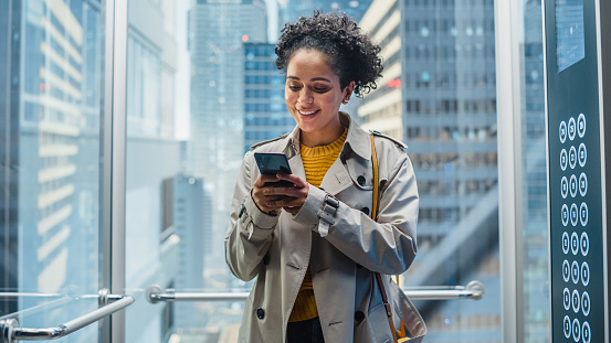 Confident Black Female Riding Glass Elevator to Office in Modern Business Center. Successful African American Manager Smile while Using Smartphone, Write Text Message, Check Social Media in a Lift.