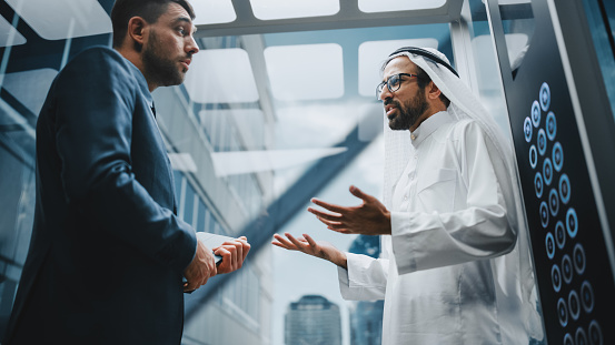 Businessman Talking with Arab Investment Partner while Riding Glass Elevator to Office in a Modern Business Center. International Corporate Associates Discussing Details of a Deal in a Lift.