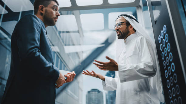 empresário conversando com parceiro de investimento árabe enquanto montava elevador de vidro para escritório em um centro de negócios moderno. associados corporativos internacionais discutindo detalhes de um acordo em um elevador. - cultura iraniana oriente médio - fotografias e filmes do acervo