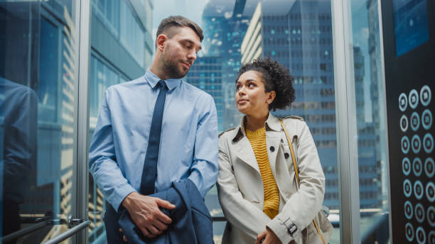 istock Two Office Colleagues Looking at Each Other While Riding in Glass Elevator in a Modern Business Center. Caucasian Male Specialist and Black Latin American Female Manager Casually Chat in the Lift. 1386884593