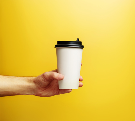A hand holds a cup for coffee on a yellow background. Takeaway drinks.
