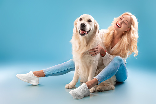 Man's Best Friend. Portrait of emotional blonde lady embracing and scratcing her happy pet, sitting on the floor with her best friend, isolated on blue studio background wall, banner, free copy space