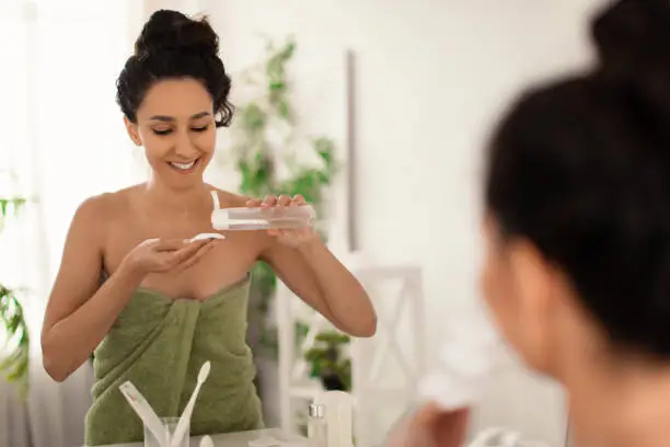 Beautiful millennial woman wearing towel after bath, refreshing her skin with lotion or tonic in front of mirror at bathroom, free space. Lovely young female enjoying facial products at home