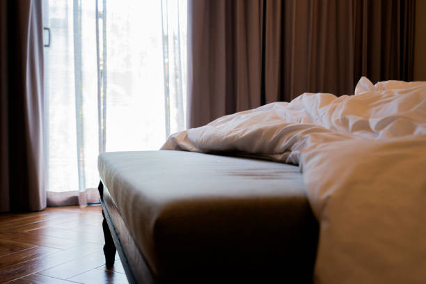 bed stool ottoman with beige fabric color upholstered stay with unmade crumpled white soft bed morning weekend sunshine light come through big window curtain home interior detail design sweet home taburete de cama otomano con tela beige color tapizado estancia con cama suave blanca arrugada sin hacer mañana luz del sol de fin de semana venir a través de la cortina de la ventana grande detalle del hogar diseño de interiores dulce hogar
