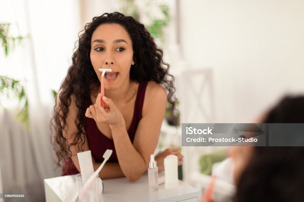 Pretty young brunette woman shaving her upper lip, removing unwanted moustache in front of mirror at home, blank space Pretty young brunette woman shaving her upper lip, removing unwanted moustache in front of mirror at home, blank space. Spa treatment, smooth skin and beauty care concept Shaving Stock Photo