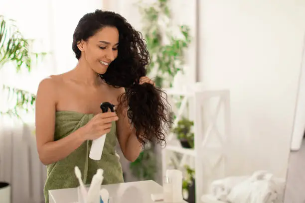 Split ends repair treatment. Smiling young woman applying haircare spray or natural essential oil on her hair near mirror at home, copy space. Beauty and domestic cosmetics concept