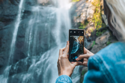 Close-up of a young female traveler photographing a waterfall with a mobile phone. First-person point of view, copy space. Concept of travel, tourism, blogging