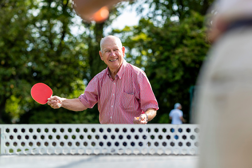 A senior man playing table tennis outdoors in a public park in Newcastle Upon Tyne, England with an unrecognisable man, who is mostly out of shot. The man is smiling while holding his table tennis racket.