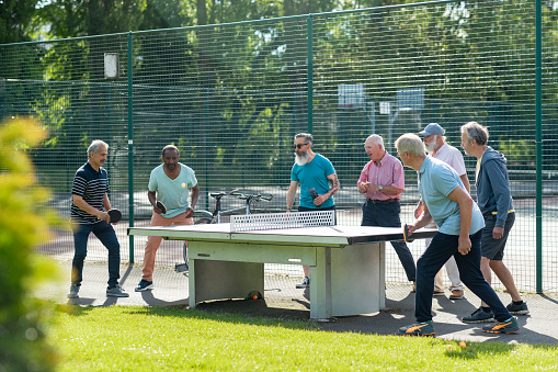 A group of senior men playing table tennis outdoors next to a tennis court in Newcastle Upon Tyne, England. Four men are playing against each other in teams of twos while the rest of the senior men watch them.