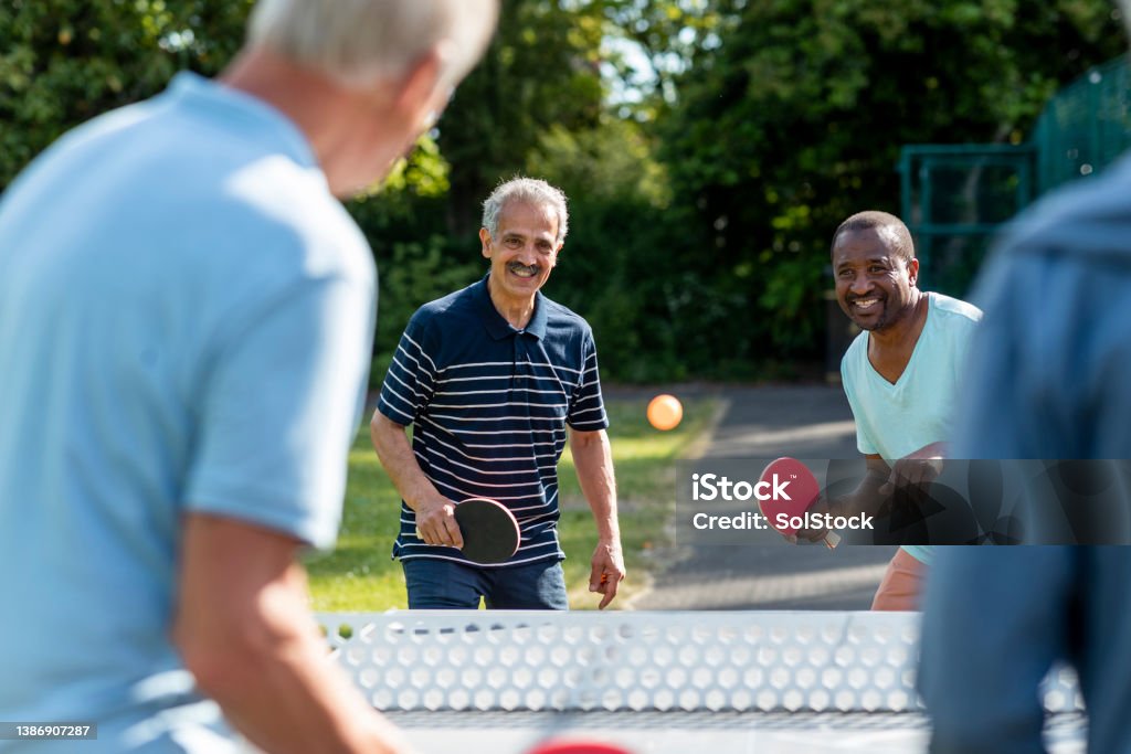 I've Got This One! A group of senior men playing table tennis outdoors in a park in Newcastle Upon Tyne, England. The main focus is two men smiling while holding table tennis rackets, getting ready to hit the ball that is approaching them. Table Tennis Stock Photo