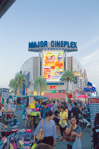 Major Cineplex cine,a building and thai people in Bangkok Rangsit at Future Park in sunset light. Screen and banner at facade of building is illuminated. People are walking on sidewalk and some market stalls are placed along both sides of building