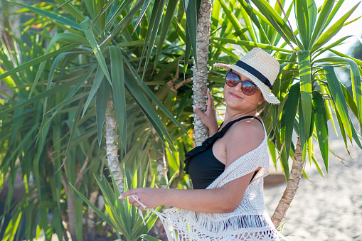 Young woman with a black swimwear, red sunglasses and sun hat in a shade of palms on a beach