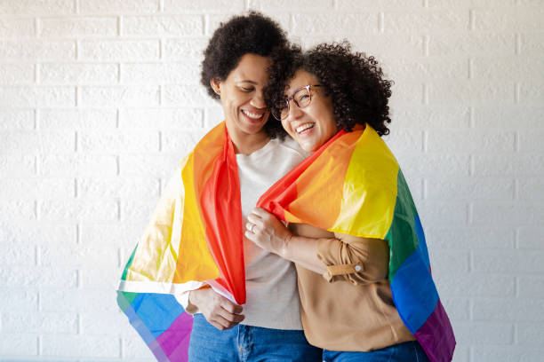 Lesbian women covered with rainbow flag Front view of lesbian couple embracing and wearing rainbow flag gay pride people stock pictures, royalty-free photos & images