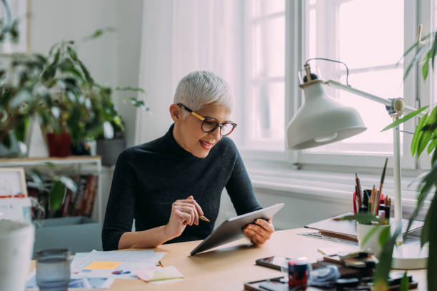 une belle femme âgée élégante et souriante regardant sa tablette tout en étant assise à son bureau dans le bureau et en travaillant - reading pad photos et images de collection