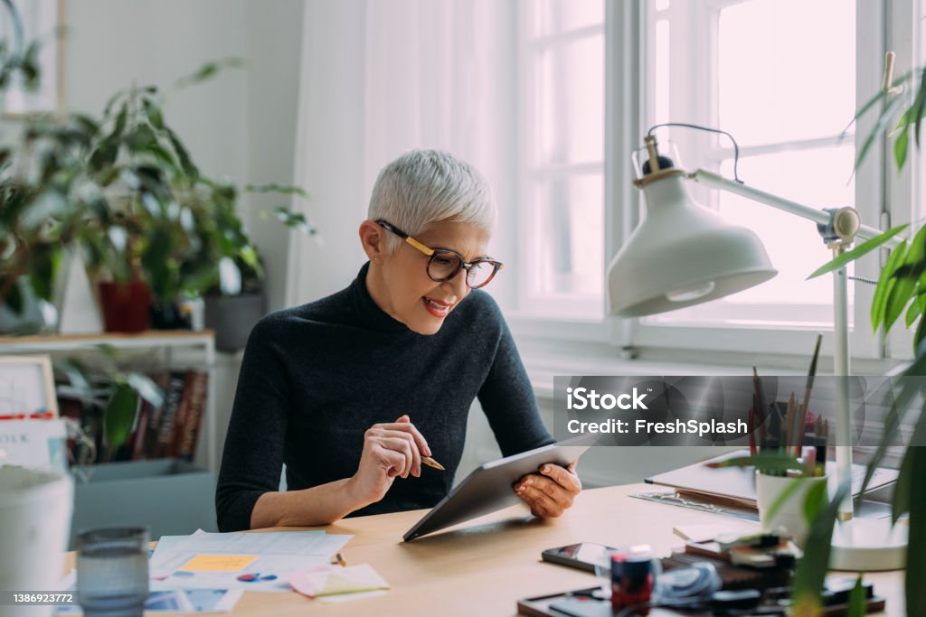 A Beautiful Smiling Elegant Senior Woman Looking At Her Tablet While Sitting At Her Desk In The Office And Working A happy charming Caucasian grey woman being focused on her work. Digital Tablet Stock Photo