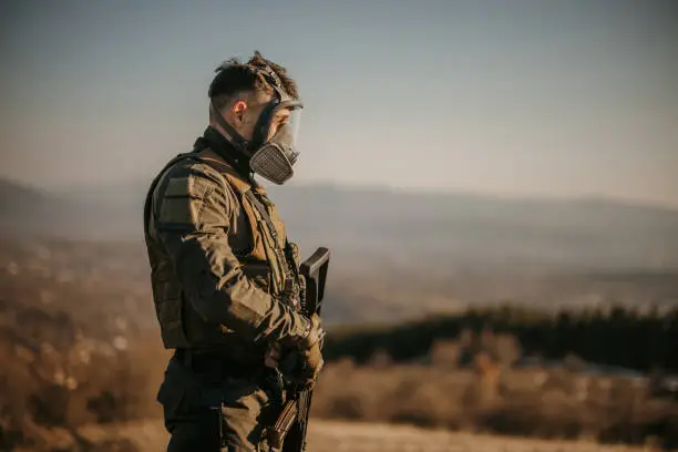 Male soldier with a rifle wearing gas mask, he is standing on a hill in nature alone.