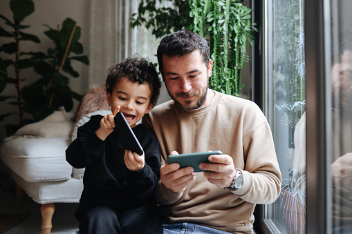 Father and son game players funs sit together at home on cozy sofa, using the tablet and gamepad