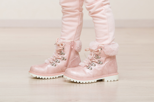 Little girl in pink trousers and leather warm boots with fluffy fur, shoelaces and zipper standing on light laminate floor at home. Closeup. Side view.