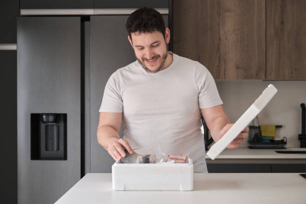 Young latin man opening his weekly delivery of fish in an EPS isothermal box. Young latin man opening his weekly delivery of fish in an EPS isothermal box, in the kitchen. polystyrene box stock pictures, royalty-free photos & images