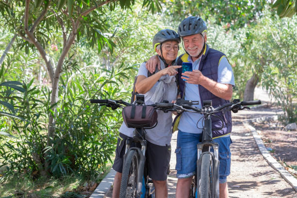 istock Active caucasian senior couple with electrobikes standing outdoors in the park looking at cellphone. Two smiling elderly people wearing helmet running in nature enjoying healthy lifestyle 1386939618