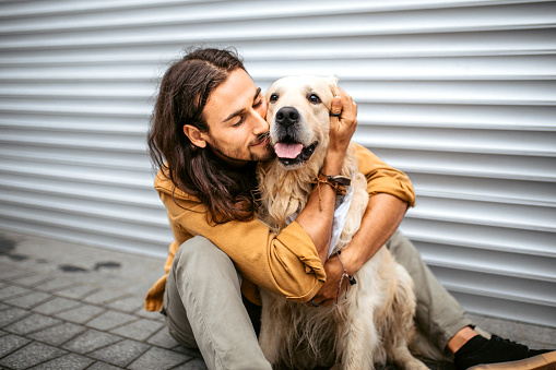 Portrait of a young handsome man hugging his golden retriever dog, sitting on a floor in a front of a white garage door.