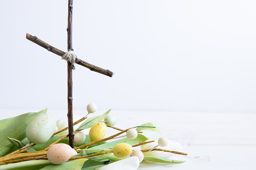 Simple Christian cross standing in a bundle of white tulips and a decorative branch of pastel coloured Easter eggs on a white background with copy space