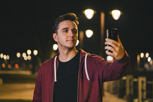 Front view portrait of one young adult caucasian man photographer standing outdoor in the city or town at night holding using mobile phone for video call or selfie photo copy space waist up