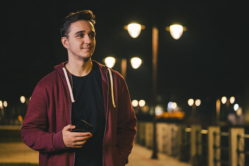 Front view portrait of one young adult caucasian man photographer standing outdoor in the city or town at night holding using mobile phone looking to the side happy smile