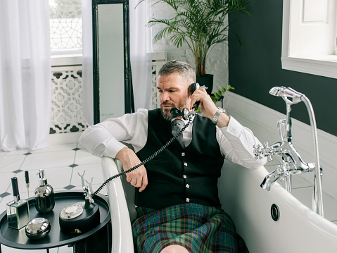 handsome mature courageous stylish man scotsman in kilt and suit talking on the phone from home. Style, fashion, lifestyle, lockdown, culture, ethnic concept.