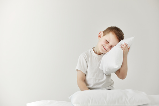 Portrait of casually dressed little boy leaning on white pillow, while hugging it, feeling tired after long full day, with cheerful smile, having rest.