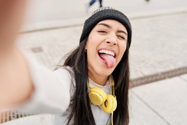 라티나는 그녀의 스마트 폰으로 그녀의 혀를 내밀고 윙크와 함께 셀카를 찍고. 셀카를 찍는 동안 재미있는 얼굴을 만드는 히스패닉 여성 - humor music headphones women 뉴스 사진 이미지