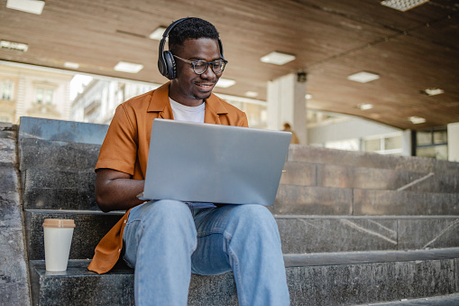 Clever African American student  typing on laptop keyboard and looking at screen while sitting on steps and working on sunny day