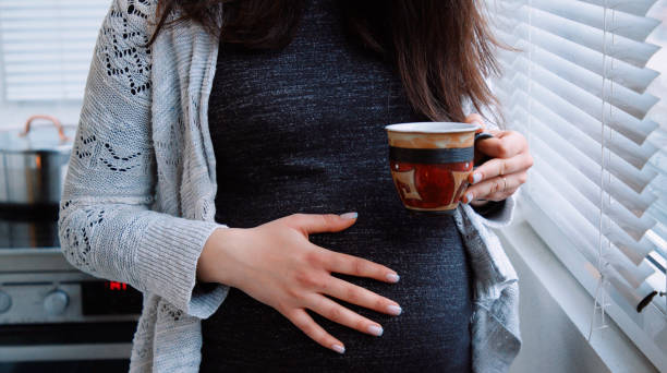 pregnant woman caresses her belly while drinking  coffee next to the window at home. - caffeine free imagens e fotografias de stock