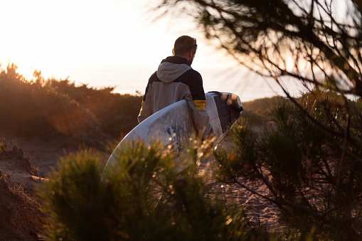 rear view of an unrecognizable young man in jacket walking at sunset by the dunes of the beach with his surfboard under his arm, leisure and hobbies concept, copy space for text