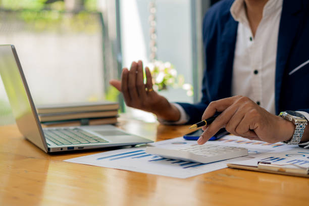 Asian businessman's hand is pressing calculator and preparing data while holding pen in finger to calculate, analyze investment advisor chart. and auditors to calculate the cost with a laptop la mano del empresario asiático está presionando la calculadora y preparando los datos mientras sostiene el bolígrafo en el dedo para calcular, analizar el gráfico del asesor de inversiones. y auditores para calcular el costo con una computadora portá