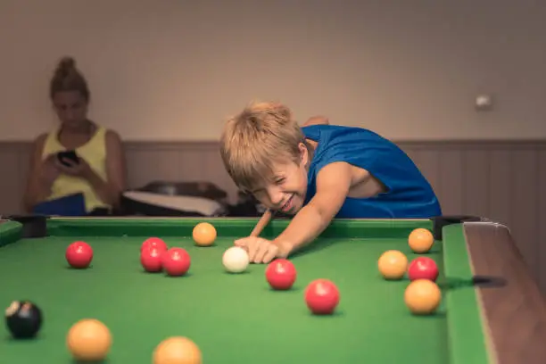 Cute boy in blue t shirt plays billiard or pool in club. Young Kid learns to play snooker. Boy with billiard cue strikes the ball on table. Active Leisure, sport, hobby concept