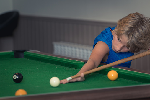 Cute boy in blue t shirt plays billiard or pool in club. Young Kid learns to play snooker. Boy with billiard cue strikes the ball on table. Active Leisure, sport, hobby concept