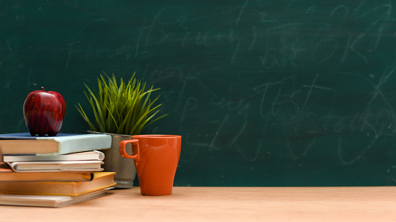 Back to school concept, Classroom study table with stack of books, mug, apple, decor plant and copy space against green chalkboard background.