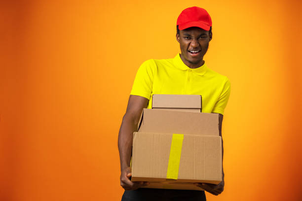 istock Smiling black male courier wearing uniform holding box in orange studio 1387011243