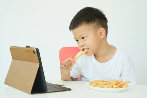 Asian boy child eating French fries while watching a video from tablet pc isolated over white wall stock photo
