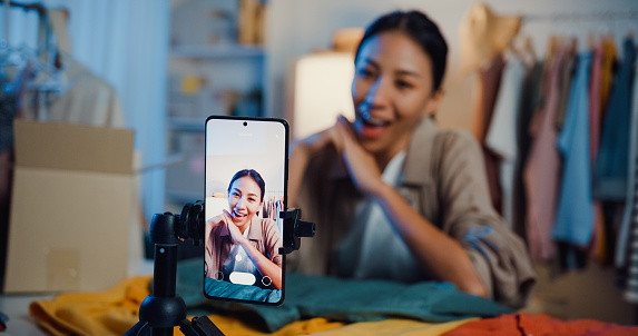 Asian woman live-streamed ecommerce sell clothes in home at night. Beautiful girl using the smartphone and tablet for recording video.