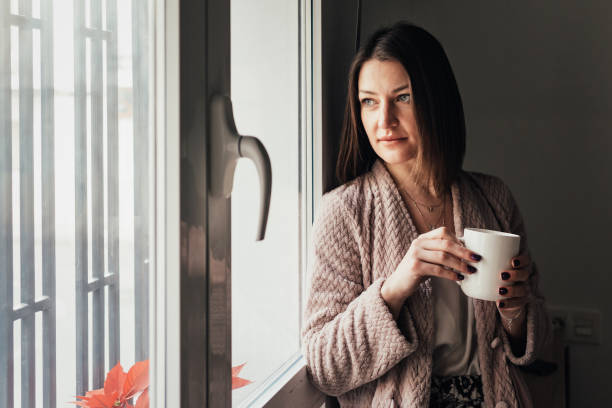 istock Relaxed woman holding a cup of coffee while looking through a window. 1387019206