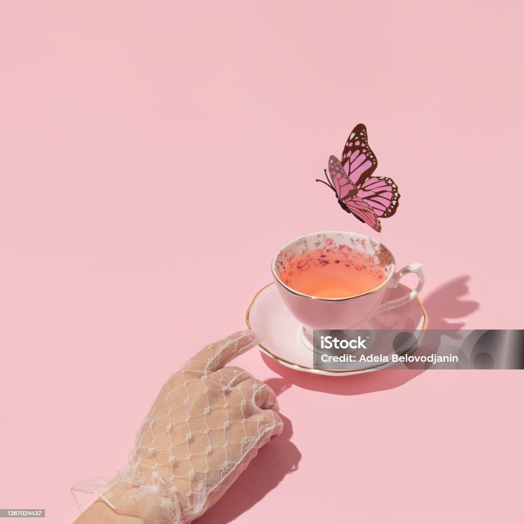 Woman hand reaching vintage cup of tea and butterfly flying above on pastel pink background. 80s, 90s retro aesthetic spring or summer concept. Minimal fashion romantic idea. Tea Cup Stock Photo