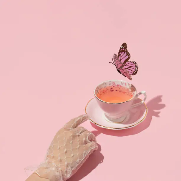 Photo of Woman hand reaching vintage cup of tea and butterfly flying above on pastel pink background. 80s, 90s retro aesthetic spring or summer concept. Minimal fashion romantic idea.