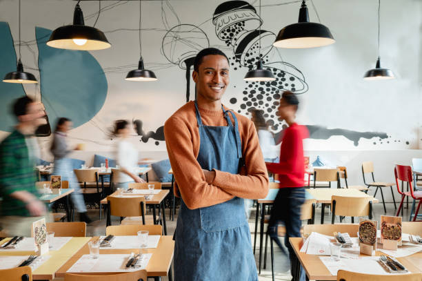 portrait of smiling male owner or waiter in cafe with unrecognizable blurred employes moving. - servitör bildbanksfoton och bilder