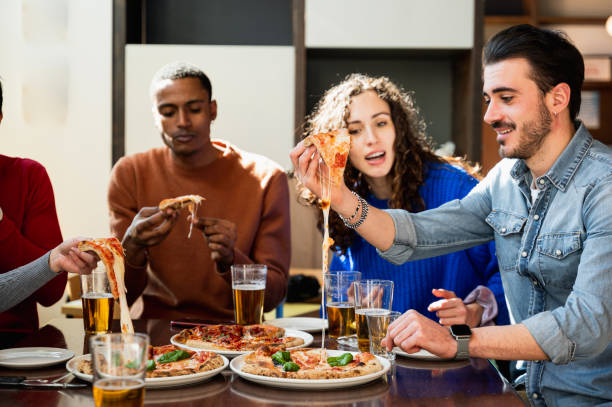 grupo multiétnico de amigos sentados à mesa comendo. os pedaços de pizza têm queijo. - stringy - fotografias e filmes do acervo