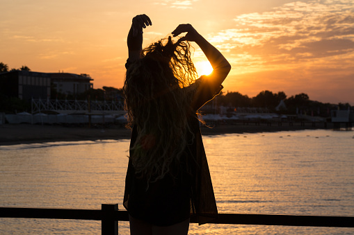 The woman stands on the sea pier of a tourist complex in Turkey. A young woman enjoys a beautiful view of the sunset over the horizon. The tourist spends his summer vacation on the Mediterranean coast. The woman is relaxing in the tourist resort of Belek.