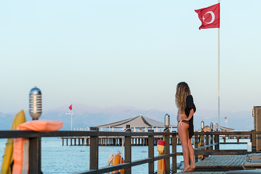 The woman stands at the edge of a sea pier. The Turetian flag is flying on the flagpole. The tourist is resting on the Mediterranean coast in Belek. Turkey.